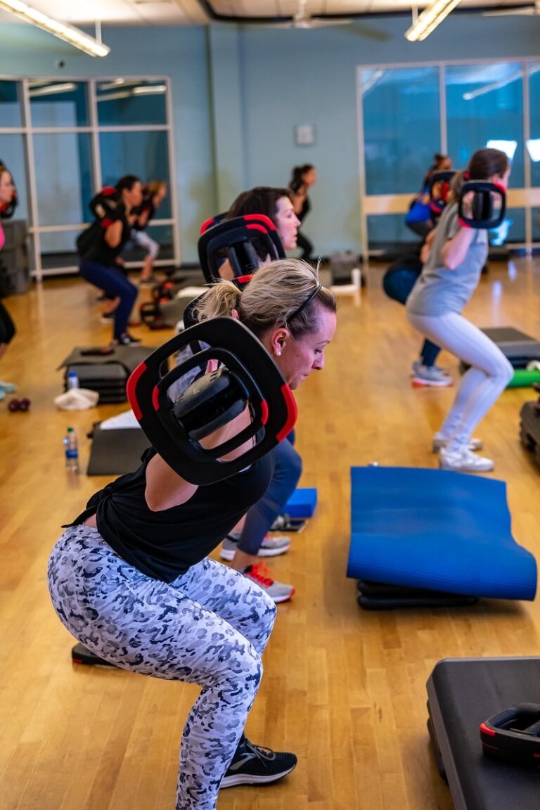 Group fitness class with participants performing squats while holding weighted bars across their shoulders in a bright studio with wooden floors and exercise mats.