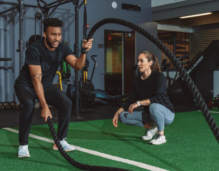 Person performing a battle rope exercise on green turf in a gym, while another person crouches nearby observing. Fitness equipment such as resistance machines and bikes are visible in the background.