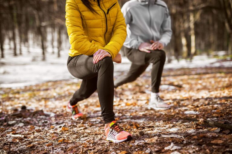 Two people performing lunges on a forest trail covered with fallen leaves, wearing athletic clothing and sneakers, with one person in a yellow jacket and the other in a gray jacket.